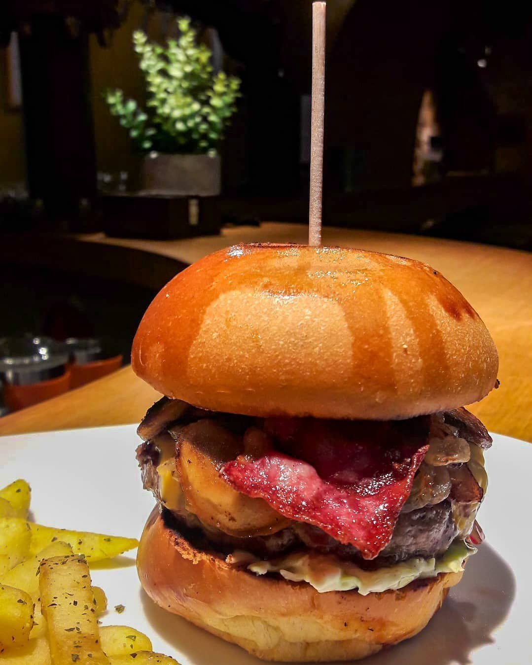 Hambúrguer em pão brioche com bacon e queijo, preso com um espeto de madeira, e batatas fritas ao lado