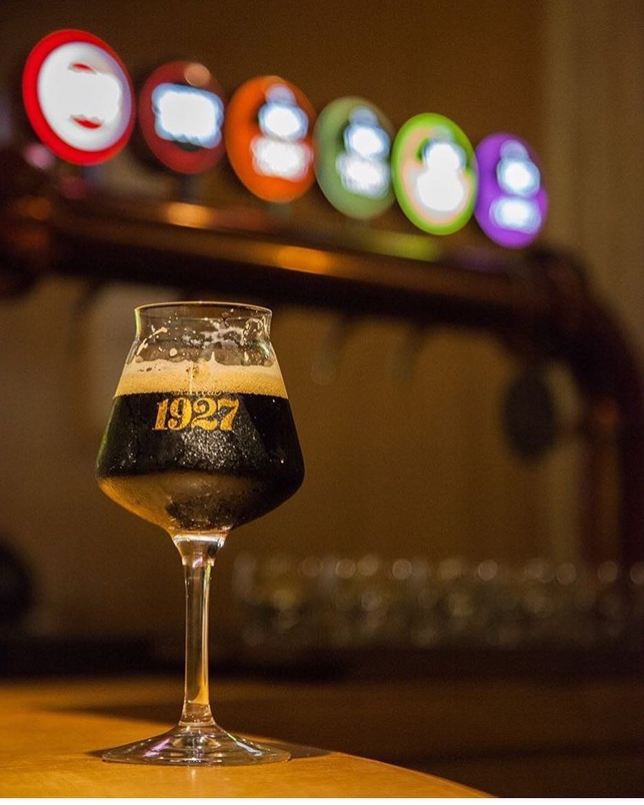 Copo de cerveja escura ao balcão, com as torneiras de cerveja iluminadas ao fundo