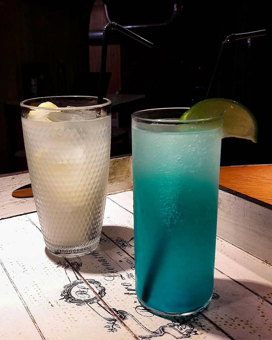 Dois copos altos com bebidas frescas, uma com rodela de lima, sobre uma mesa de madeira